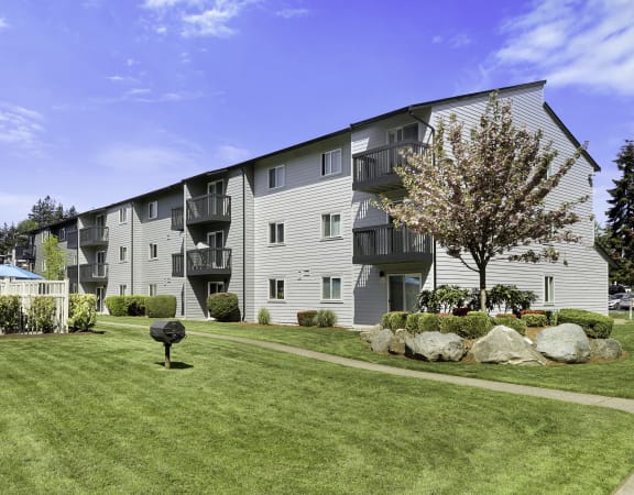 a grassy area with a tree in front of an apartment building at Pacific Park Apartment Homes, Edmonds, WA