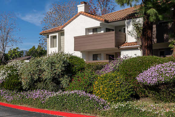 A white house with a red roof and a brown awning is surrounded by a garden with purple flowers.