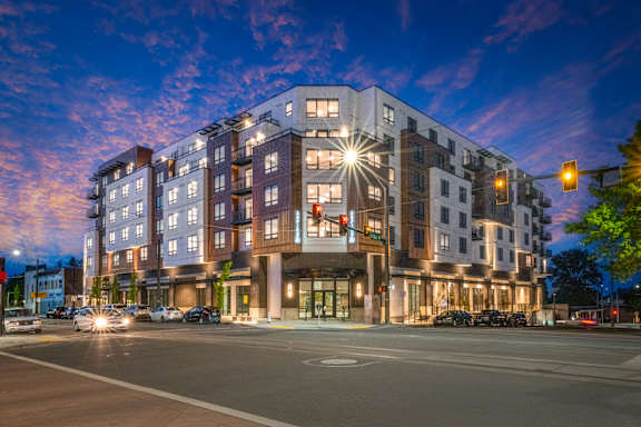 A modern apartment building is lit up at dusk with cars parked in front.