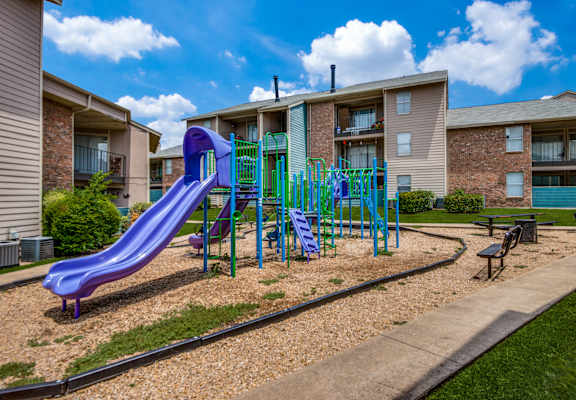 A purple slide in a playground in front of apartment buildings.