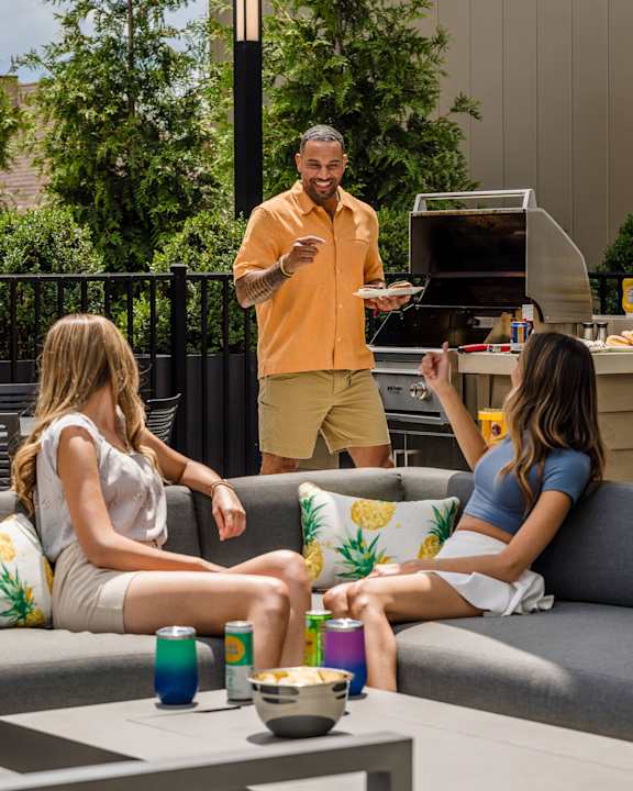 A man in an orange shirt is grilling food for two women sitting on a couch.