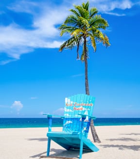 A blue beach chair with the words "Holiday Inspiration Book" written on it is placed on a sandy beach with a palm tree in the background.