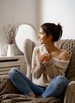 A woman in a beige sweater and blue jeans sits on a couch with her hands clasped together.