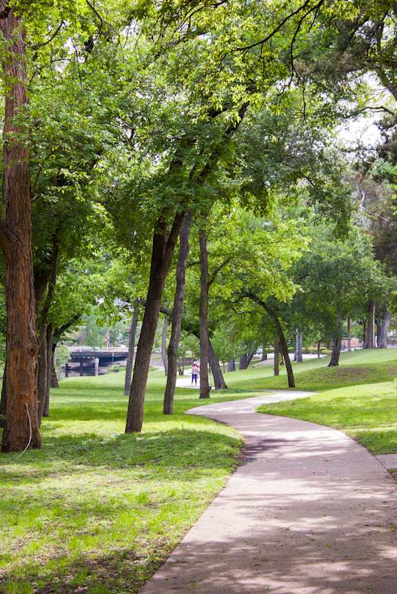 a path through a park with trees and a bench at Aero White Rock, Dallas, 75238