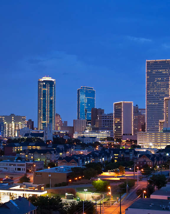 A city skyline at dusk with buildings illuminated against a darkening sky.