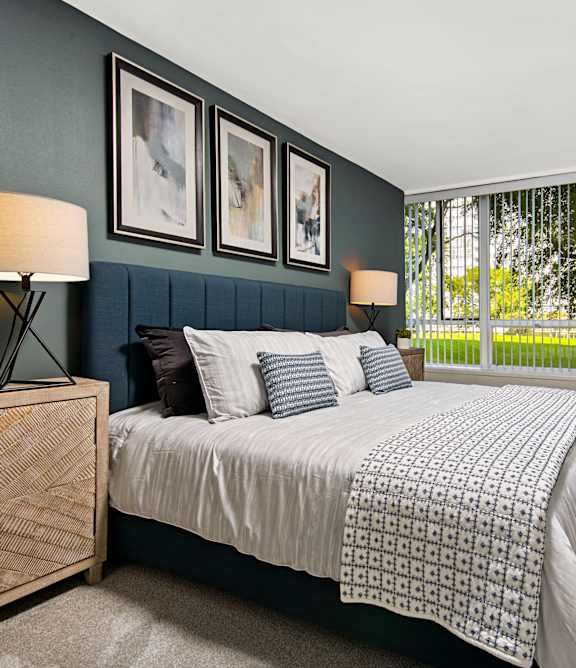 A bedroom with a bed, nightstand, and a window with a view of trees.