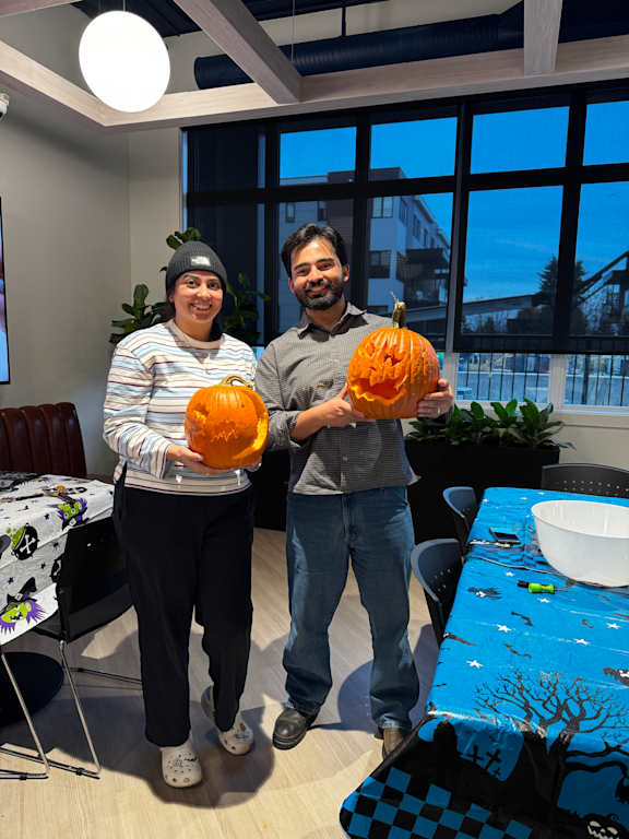 Two people holding pumpkins in a room with a table and chairs.
