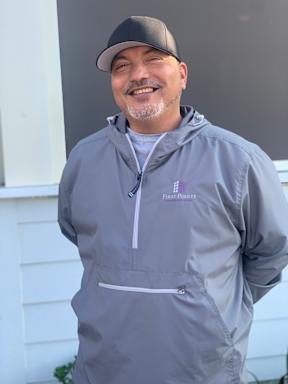 A man wearing a grey jacket and a cap is smiling.
