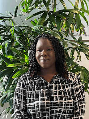 A woman with curly hair and a plaid shirt stands in front of a green plant.