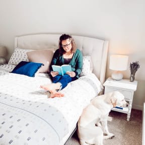 a woman sitting on a bed reading a book with her dog
