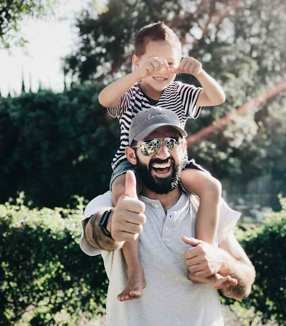 A man is giving a thumbs up while holding a child on his shoulders.