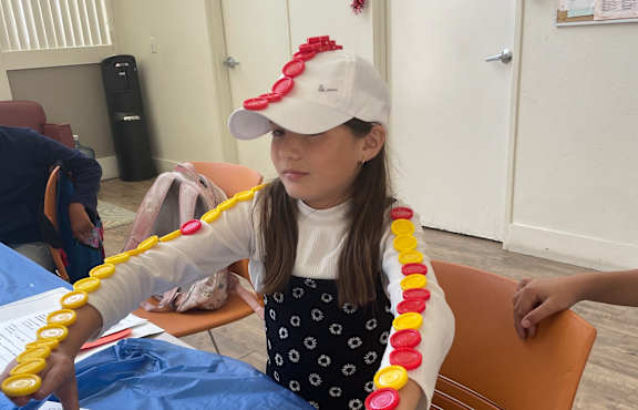 A woman is sitting in a chair with a white hat on and a yellow and red striped scarf around her neck.