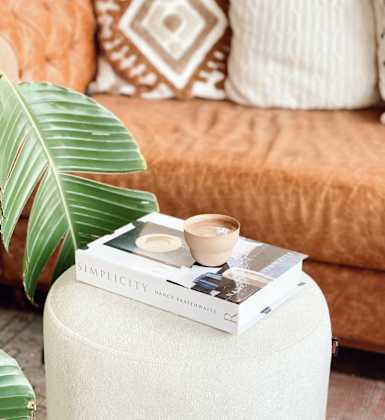 A book titled "SIMPLICITY" by Nancy Drew Haines is placed on a white stool with a green leaf and a cup of coffee beside it.