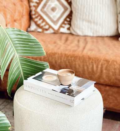 A book titled "SIMPLICITY" by Nancy Drew Haines is placed on a white stool with a green leaf and a cup of coffee beside it.