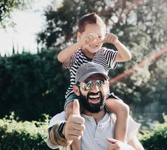 A man is giving a thumbs up while holding a child on his shoulders.