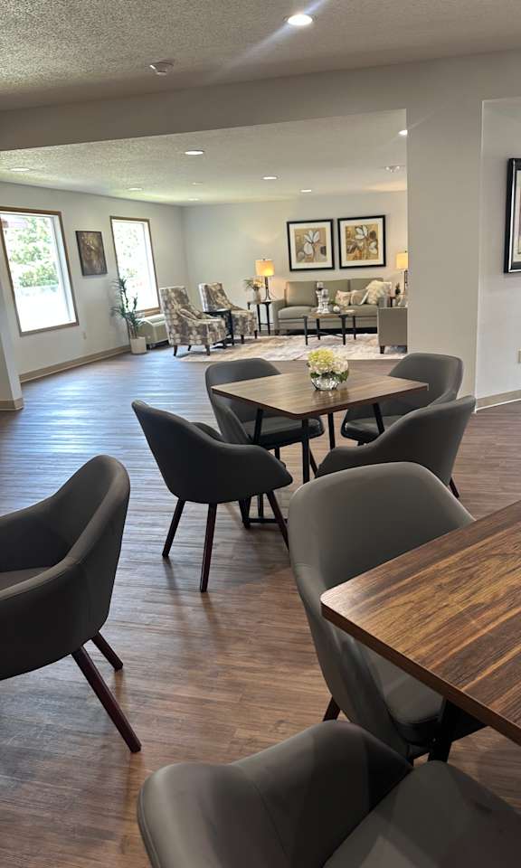 A long wooden table is surrounded by grey chairs in a room with a white wall and a window.