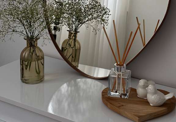 A round mirror on a white shelf with a vase of dried flowers and a diffuser with reed sticks.