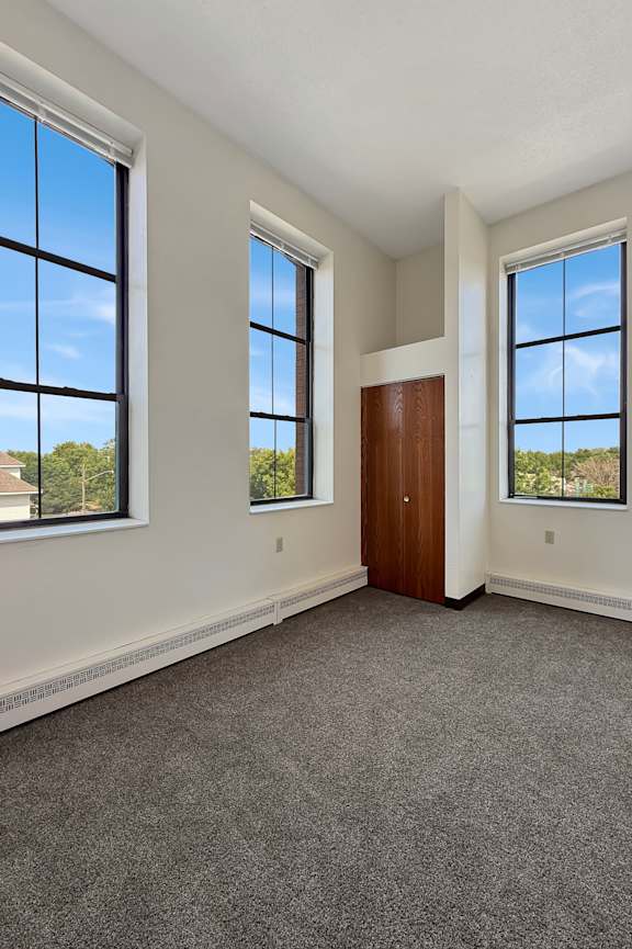 A room with a carpeted floor, a brown door, and two windows.