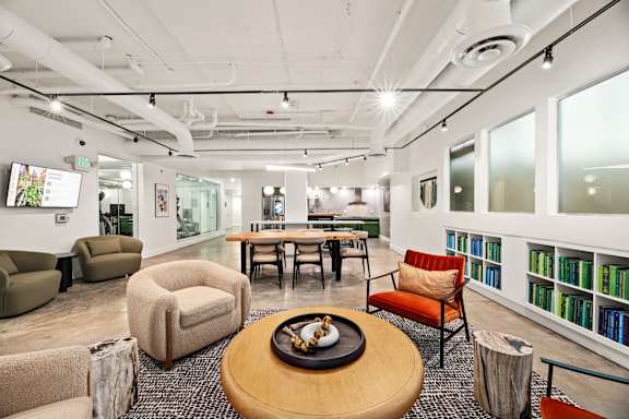 A modern office space with a wooden table in the center and a bookshelf filled with books on the right.