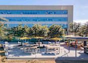 Thumbnail 22 of 24 - A large building with a glass facade is surrounded by trees and has an outdoor seating area with tables and chairs.