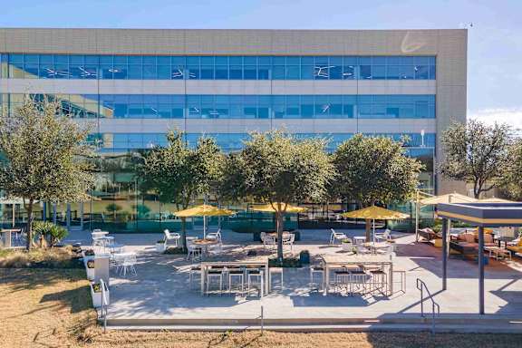 A large building with a glass facade is surrounded by trees and has an outdoor seating area with tables and chairs.