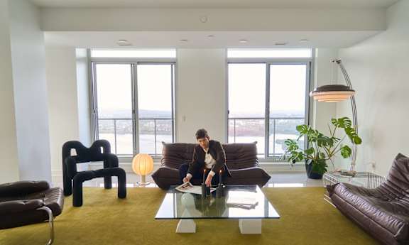 A round wooden table with a bowl on it is in the center of a room with a large window.