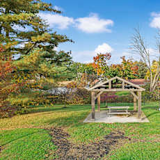 A picnic area with a bench and a pavilion is surrounded by trees with autumn foliage.
