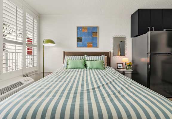 A bed with a blue and white striped comforter sits in a room with a painting on the wall and a refrigerator to the right.at The Teale New Haven Apartments, Melbourne, FL, 32904