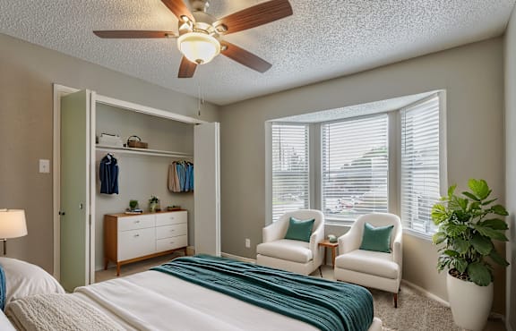 A bedroom with a bed, two chairs, a ceiling fan, and a potted plant.