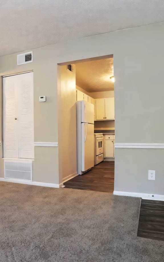 an empty living room with a door to the kitchen and a hallway with a refrigerator