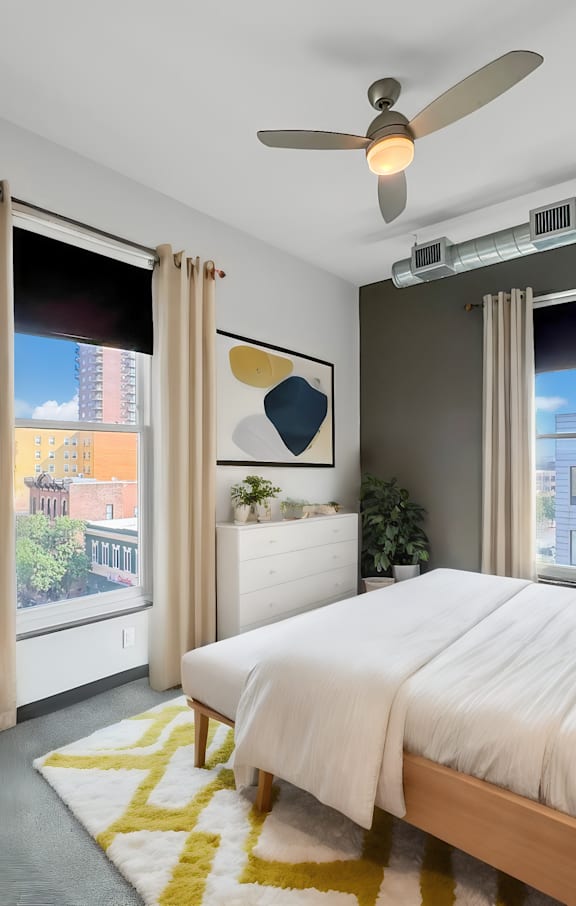 A bedroom with a large bed, a ceiling fan, and a view of a cityscape through the windows.