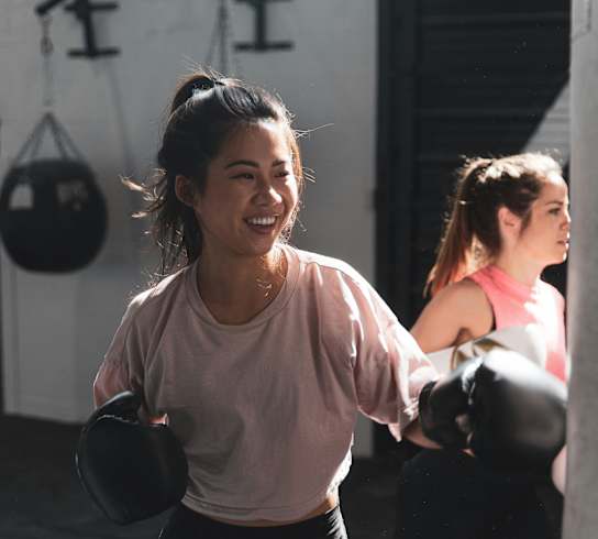 Two women in a gym, one wearing a pink shirt and black pants, the other wearing a pink tank top and black leggings, both wearing boxing gloves.