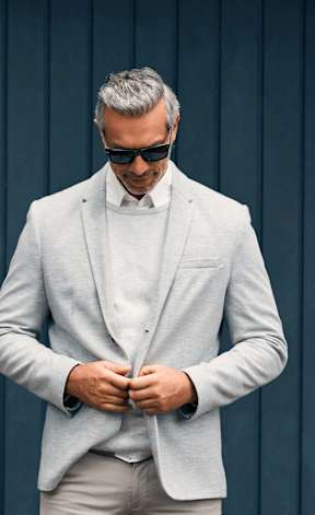 A man in a light grey suit and sunglasses is adjusting his jacket.