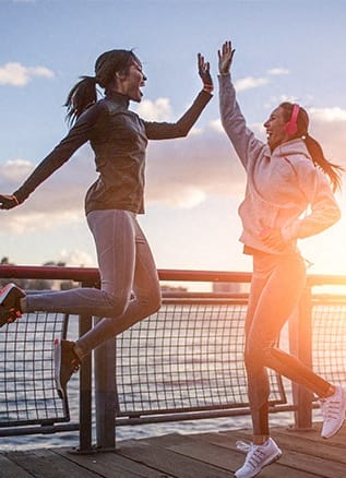 2 women running along a river at sunset & doing a high-five