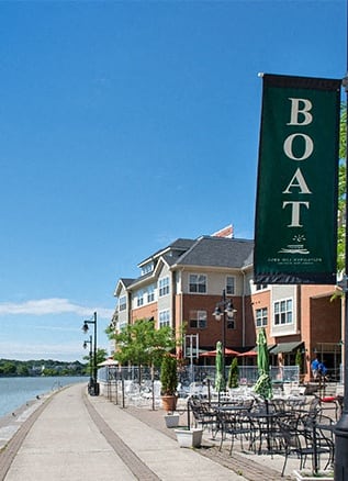 Exterior view of Corn Hill Landing from the Genesee Riverway Trail along the Genesee River showing restaurant tables & chairs, lamp posts and apartments overlooking the river