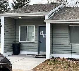 A small house with a grey door and a black trash can in front.
