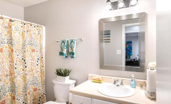 A bathroom with a white toilet, sink, and a mirror reflecting a room with a bed.