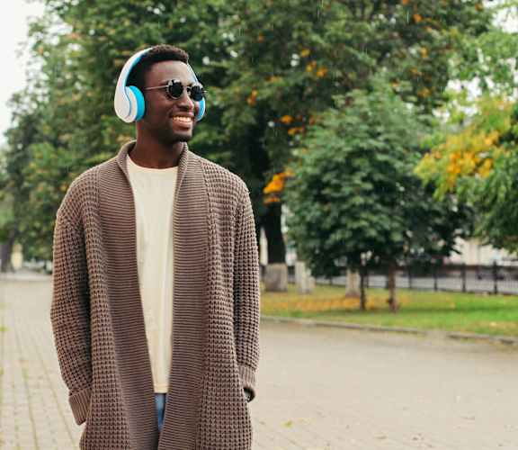 A man in a brown coat and sunglasses is wearing headphones and standing on a sidewalk.