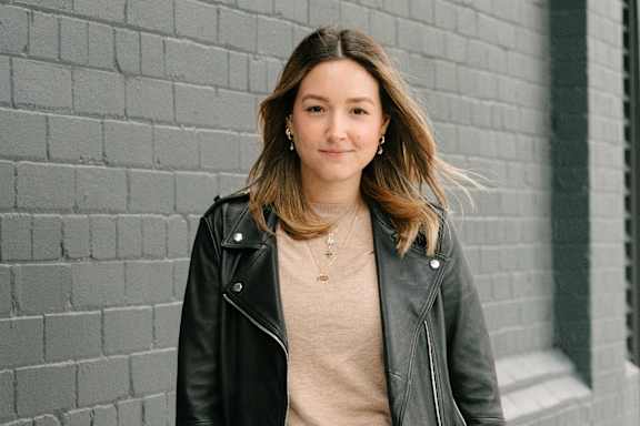 A woman with a black leather jacket and a tan shirt stands in front of a grey brick wall.