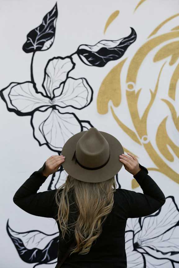A woman with a hat standing in front of a wall with a drawing of leaves.