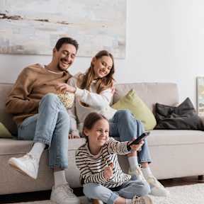 A family of three is sitting on a couch with a child in the middle holding a remote control.