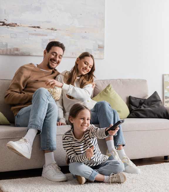 A family of three is sitting on a couch with a child in the middle holding a remote control.