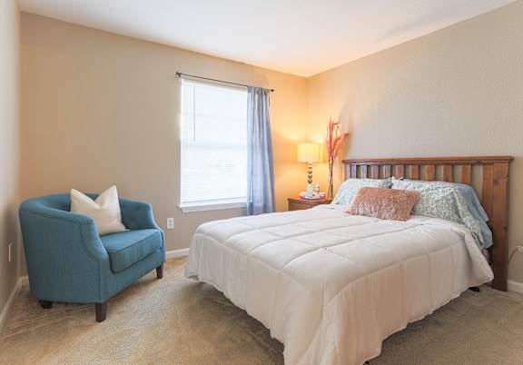Bright, spacious bedroom with white bed and blue accent chair in an apartment home at Waterbrook Apartments in Lincoln, Nebraska, professionally operated by Highlands Vista Group.