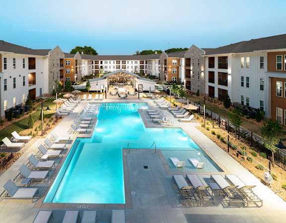 A large swimming pool surrounded by sun loungers and chairs in front of apartment buildings.