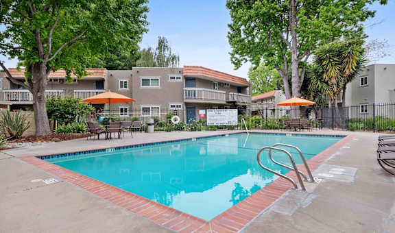Pet-Friendly Apartments in Hayward, CA - Glen Oaks - Outdoor Rectangular Pool with Poolside Lounge Seating, Tables with Orange Umbrellas and Chairs, and Exterior Views of the Property Buildings.