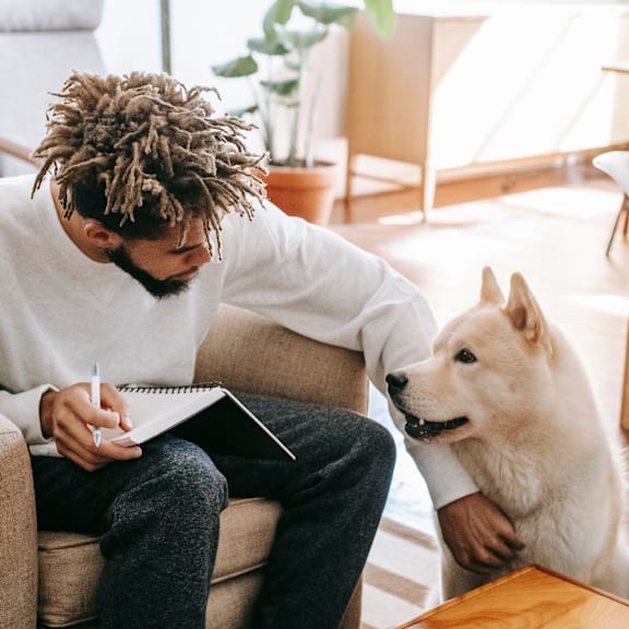 a man sitting on a couch with a dog and writing in a notebook