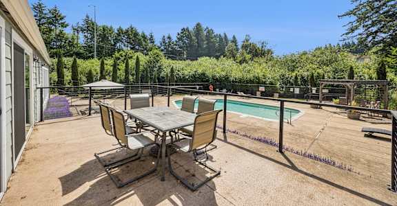 A table and chairs are set up on a patio with a pool and trees in the background.