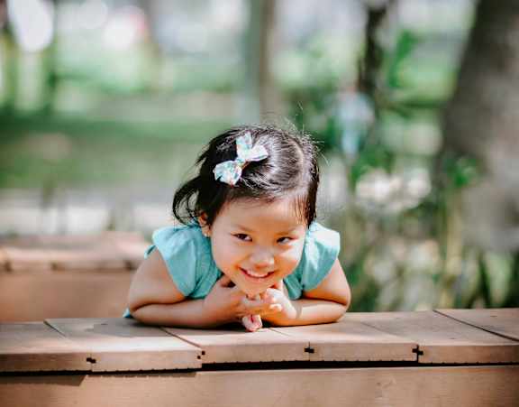 A little girl with a white bow in her hair is leaning on a wooden structure.