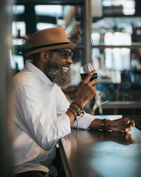 A man in a hat and glasses is sitting at a table with a glass of wine in his hand.
