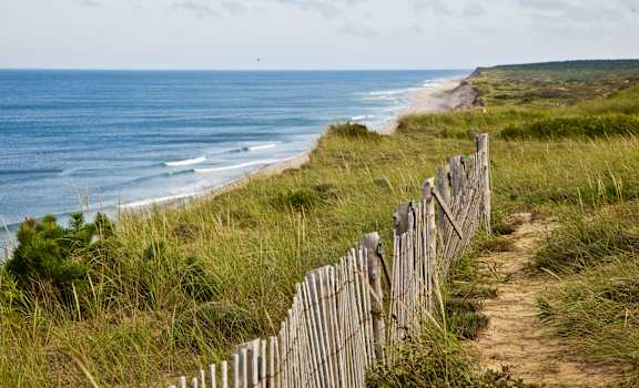 A wooden fence runs along a grassy path next to the ocean.